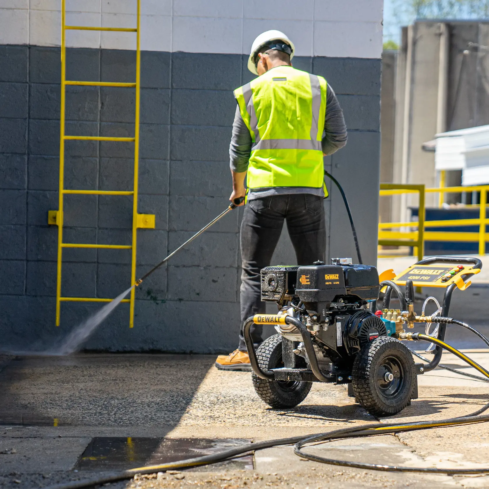 A person wearing a high-visibility vest and helmet uses a DEWALT DXPW60606 pressure washer to clean a concrete surface outdoors. A yellow ladder is attached to the wall nearby.