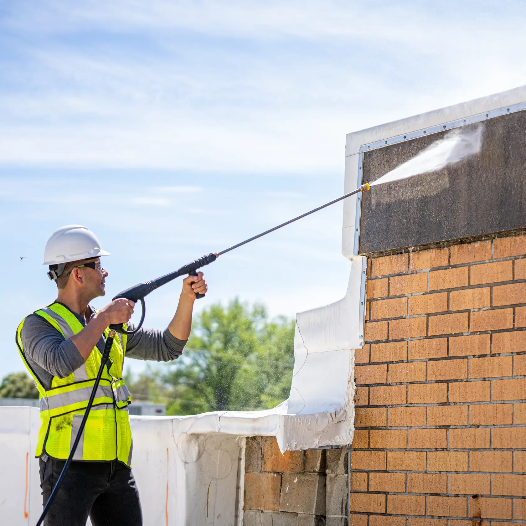 Person wearing a safety helmet and reflective vest using a DEWALT pressure washer to clean the exterior wall of a building.
