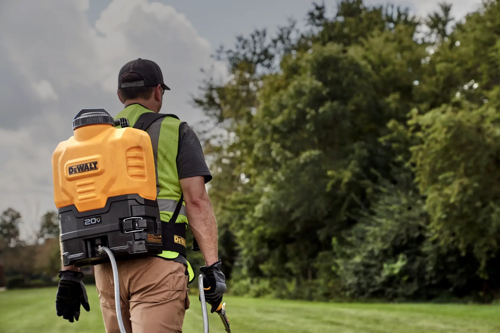 A person wearing a DEWALT 20V backpack sprayer, walking outdoors on grass near trees. The individual is dressed in work clothes and gloves, with the sprayer visible on their back.