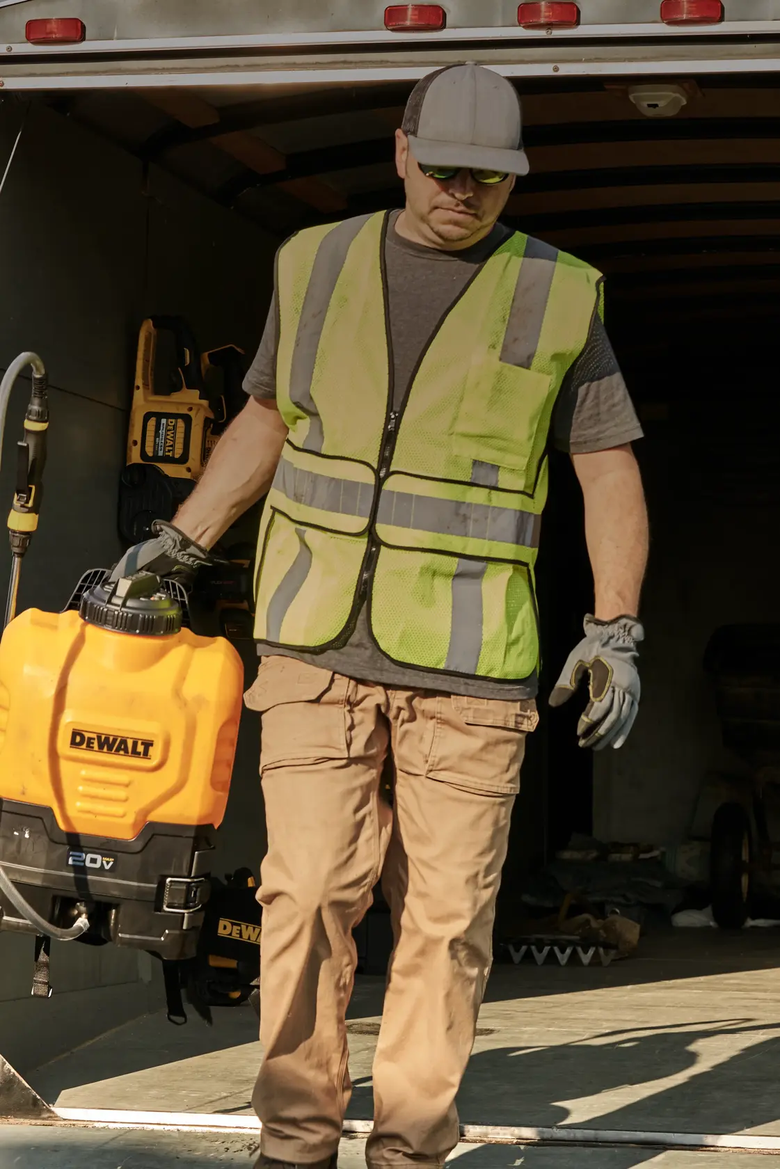 A person wearing a safety vest and gloves is carrying a DEWALT 20V sprayer, walking out of a trailer.
