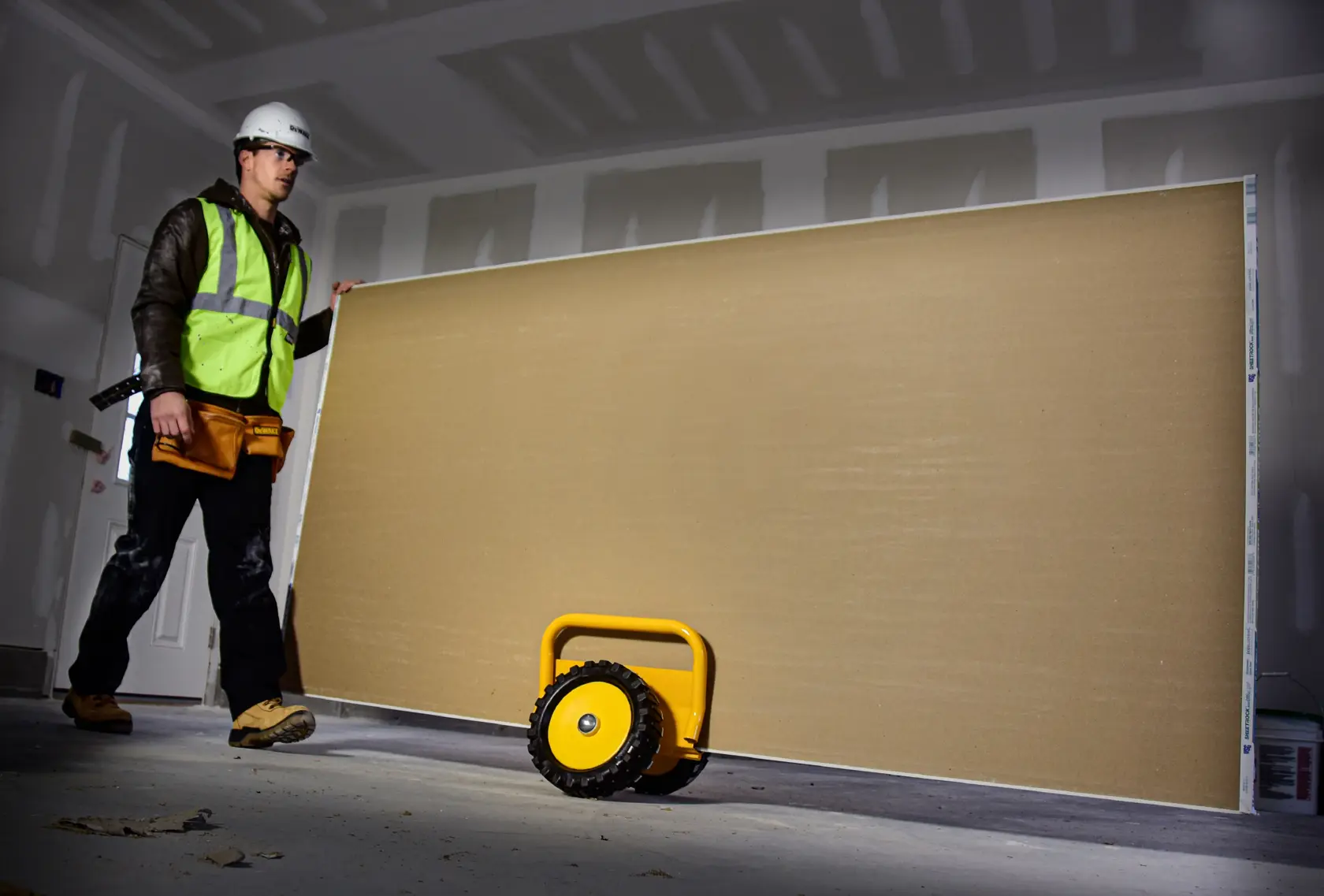 A construction worker wearing safety gear stands in a partially finished room next to a large drywall sheet being transported with a yellow DEWALT panel cart.