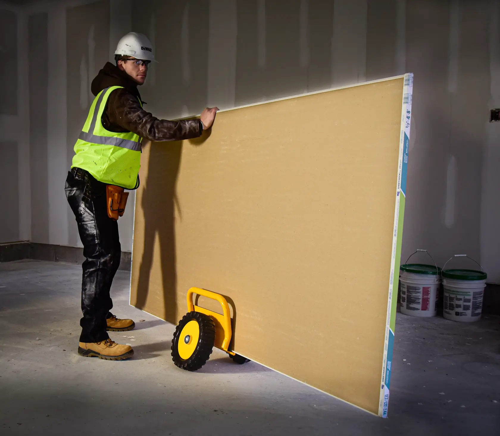 A construction worker wearing a yellow safety vest and hard hat is using a DEWALT DXWT-PS200 panel trolley to transport a large drywall sheet inside an unfinished room.