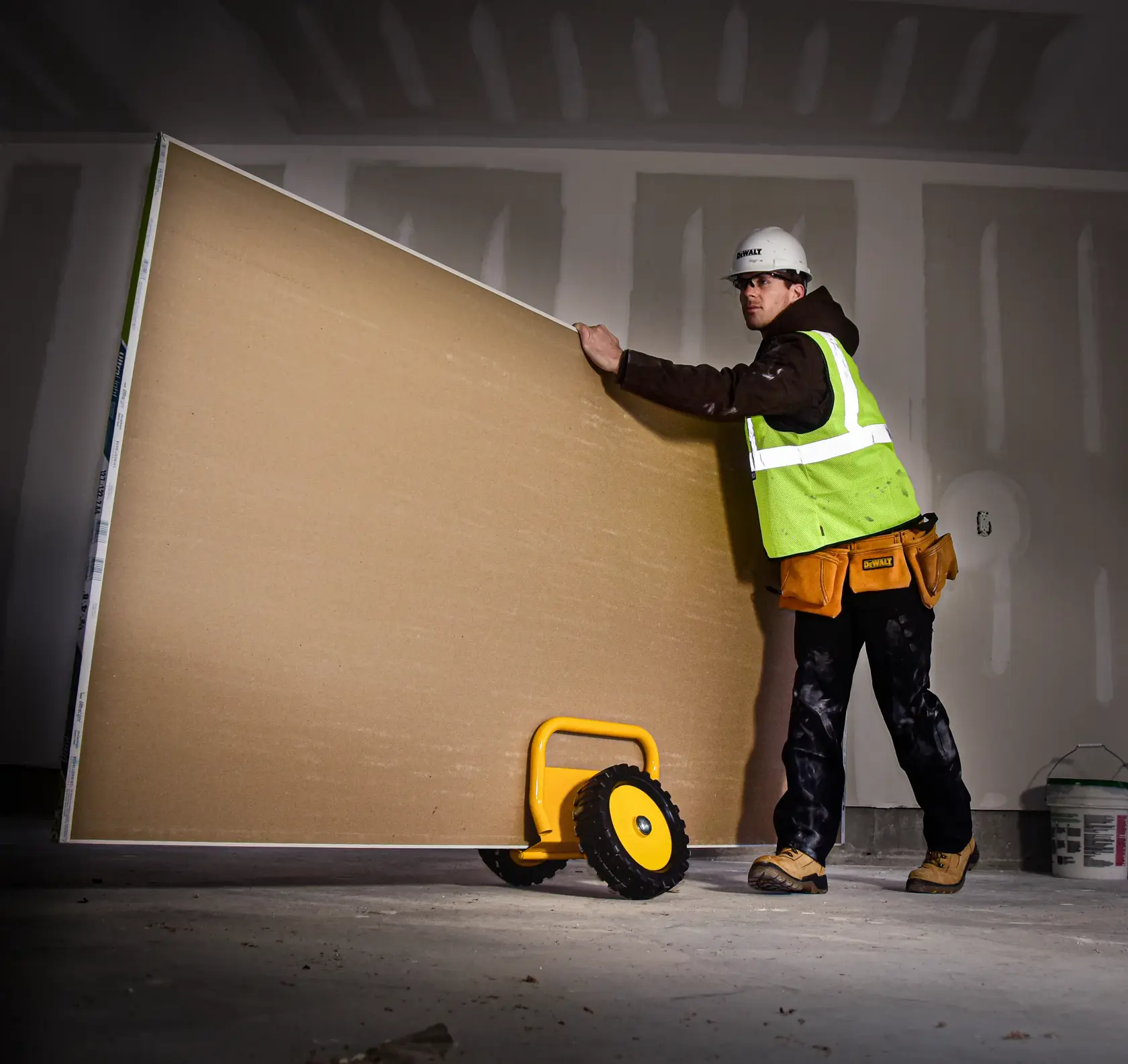 A construction worker using a yellow DEWALT drywall panel carrier with wheels to transport a large sheet of drywall in an unfinished room. The worker is wearing a safety vest, tool belt, hard hat, and work boots.