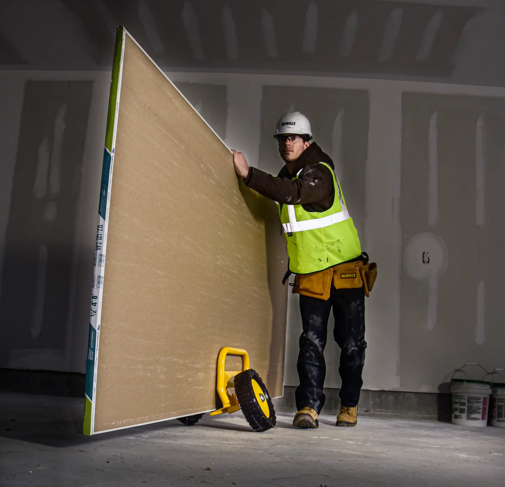 A construction worker in safety gear uses a DEWALT drywall panel dolly (DXWT-PS201) to move a large sheet of drywall inside a building under construction.